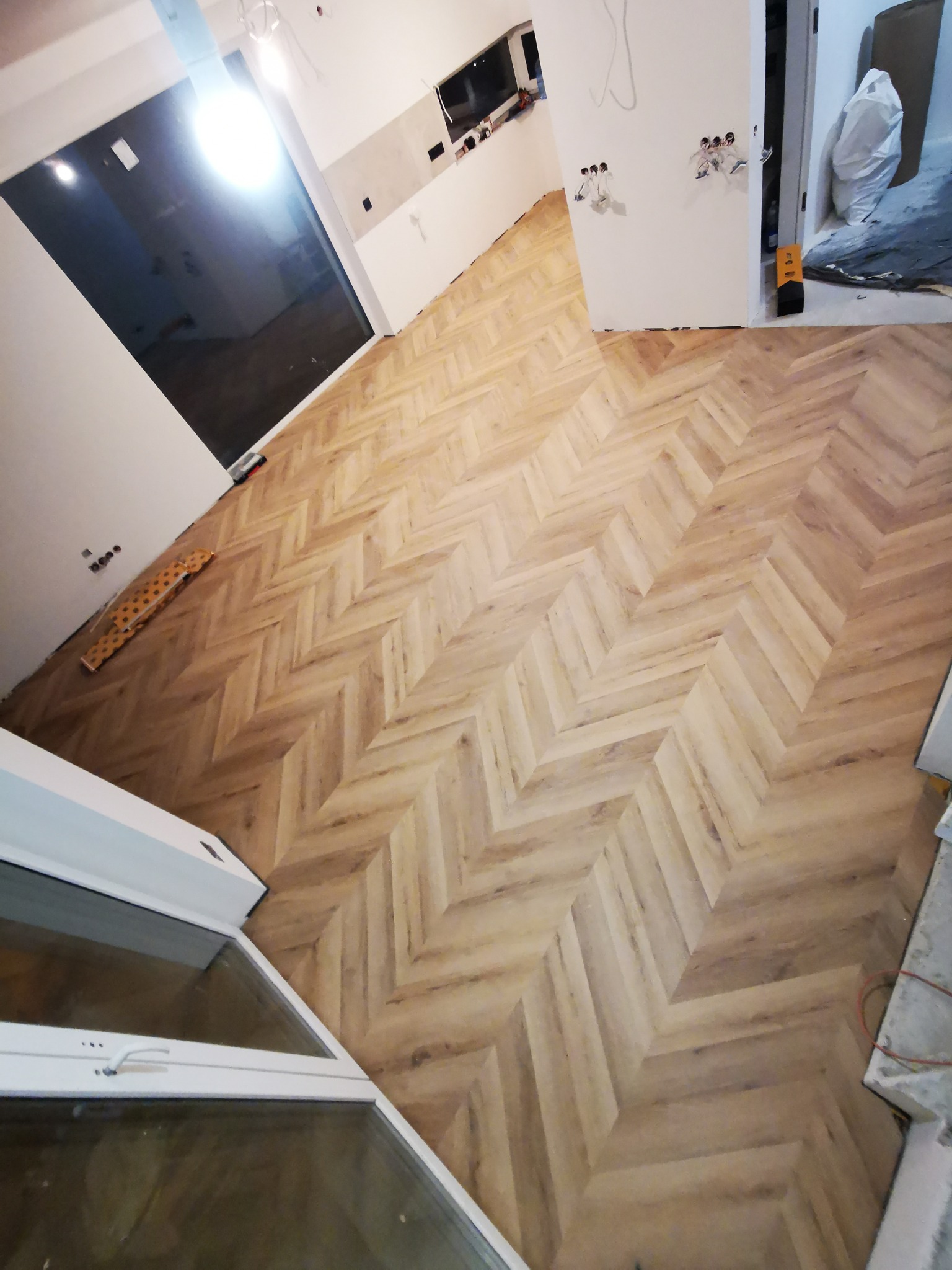 Jasnobrązowa podłoga ułożona w jodełkę francuską, widok z góry na wykończone wnętrze z białymi ścianami i dużym oknem.