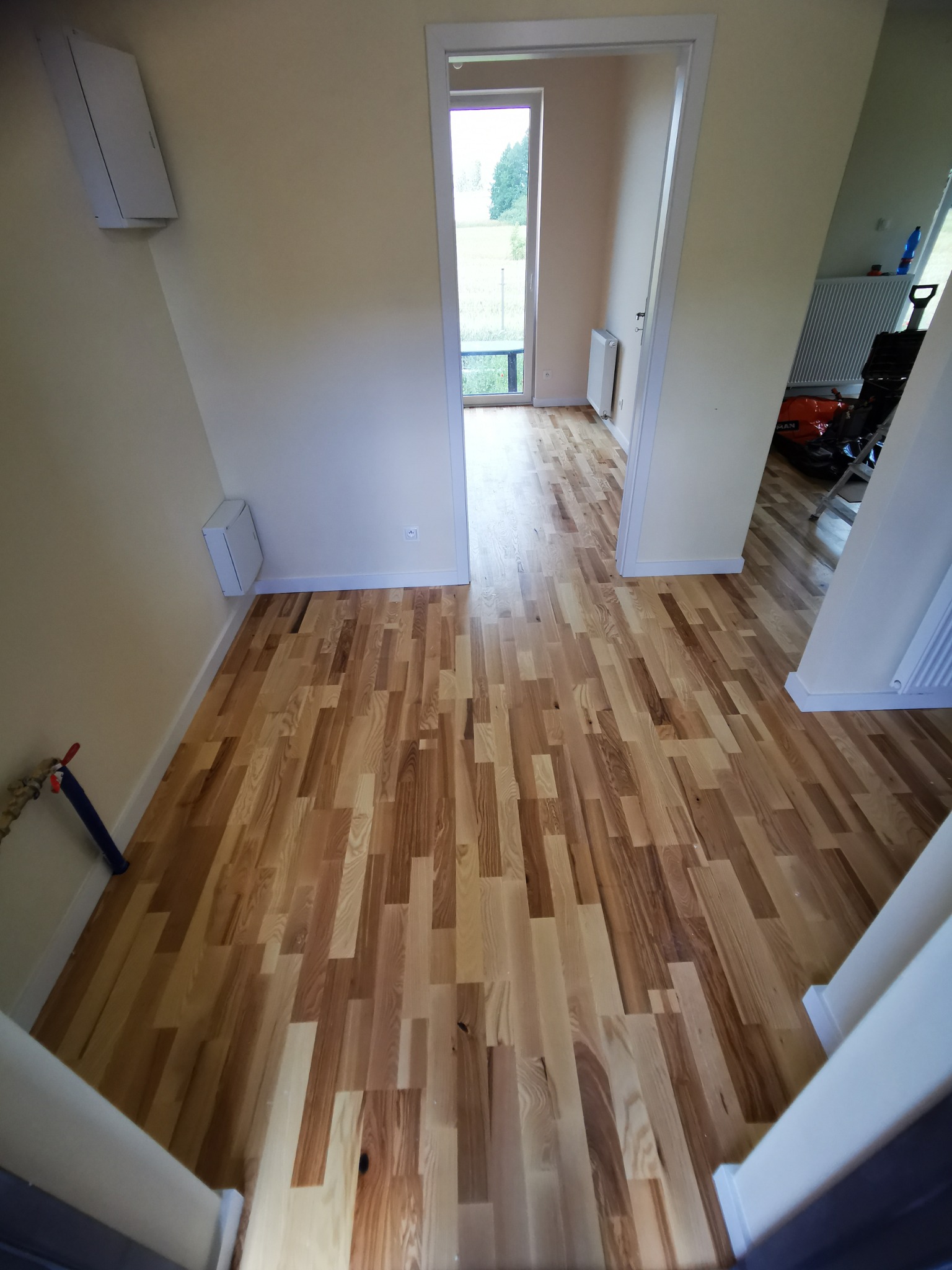 Jasna podłoga z drewnianych desek ułożona w korytarzu. Widoczne przejście do pokoju z oknem. Na ścianie biała skrzynka. U dołu fragment schodów.