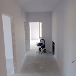 Wnętrze po szpachlowaniu, z maszyną do natrysku. Ściany gładkie, przygotowane do malowania. Widoczne surowe ościeżnice i instalacja w ścianie.