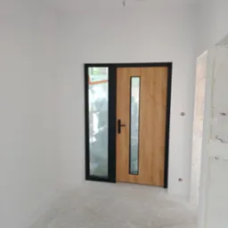 Wnętrze w trakcie wykańczania: białe ściany, drewniane drzwi z czarną ramą i przeszkleniem. Podłoga szpachlowana, widoczne ślady prac wykończeniowych.