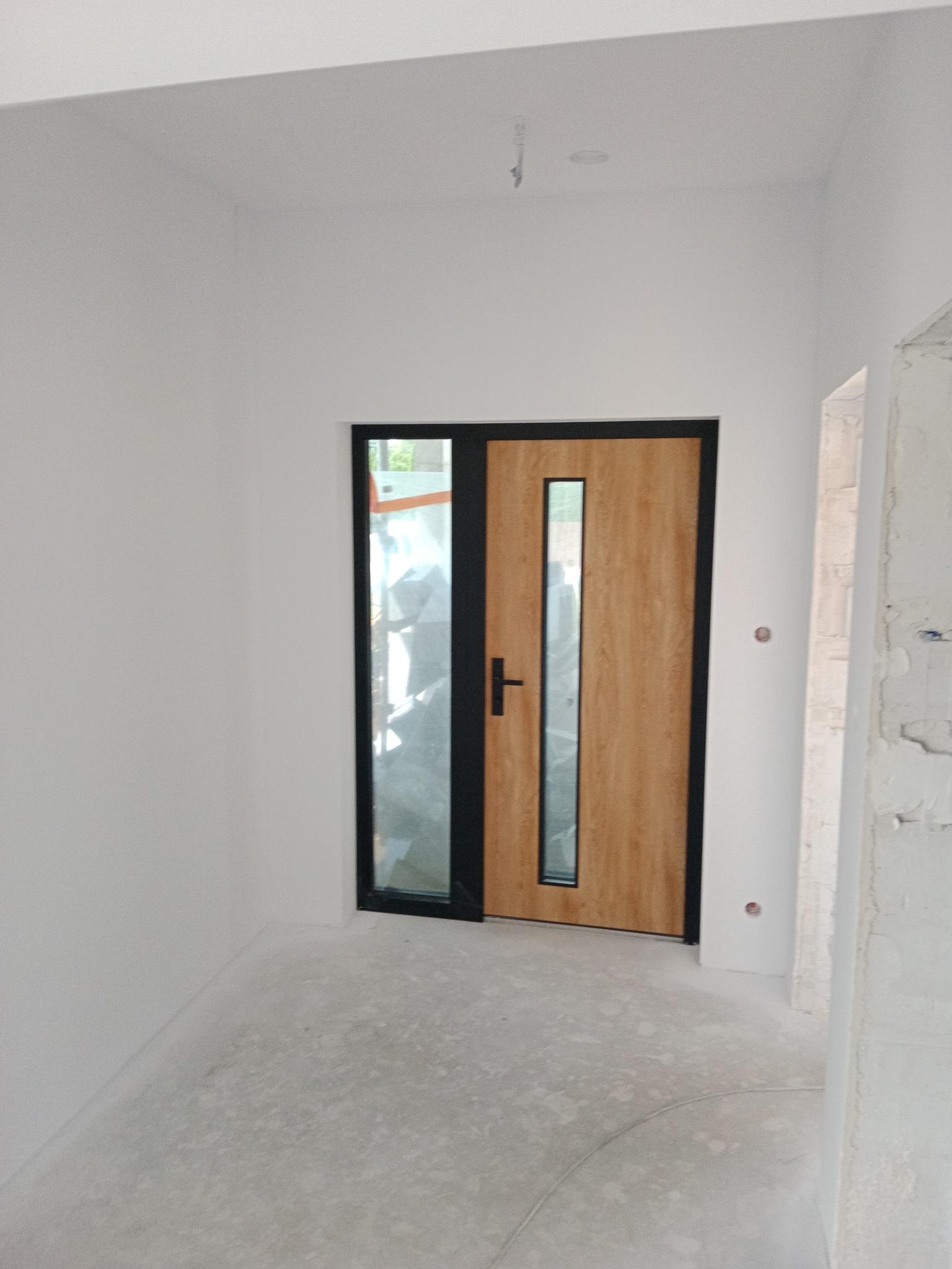 Wnętrze w trakcie wykańczania: białe ściany, drewniane drzwi z czarną ramą i przeszkleniem. Podłoga szpachlowana, widoczne ślady prac wykończeniowych.