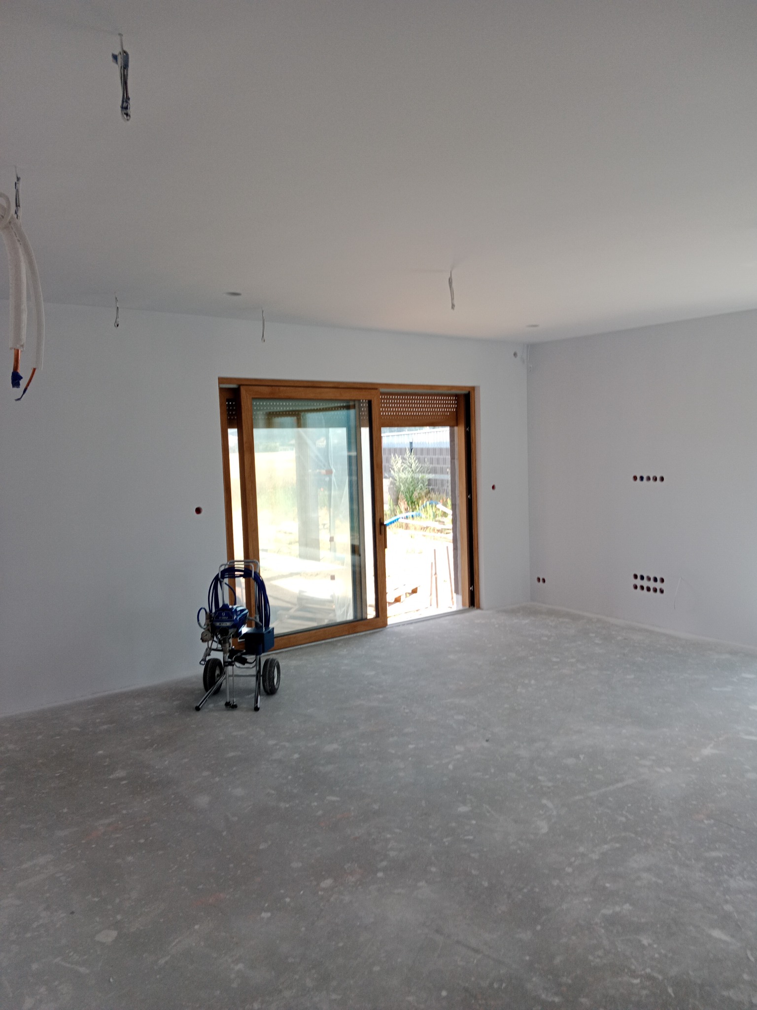 Wnętrze po malowaniu na biało, widoczny agregat malarski przy oknie balkonowym. Ściany świeżo pomalowane, sufit również biały, widoczne punkty oświetleniowe.