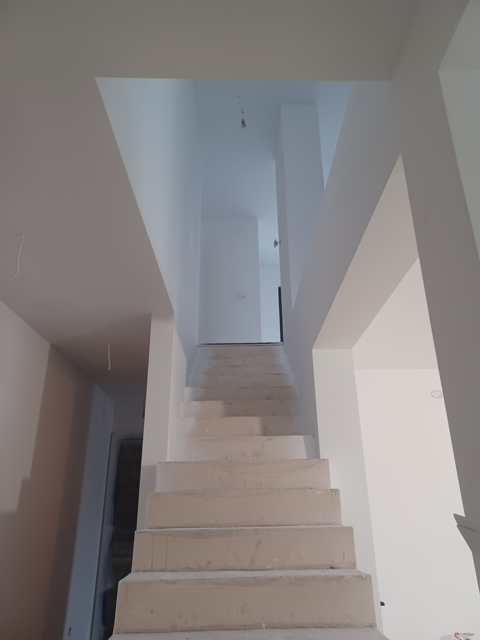 Wnętrze domu w trakcie wykańczania: surowe schody prowadzą na górę, ściany i sufit świeżo pomalowane na biało, widoczne niedoskonałości wymagające szpachlowania.