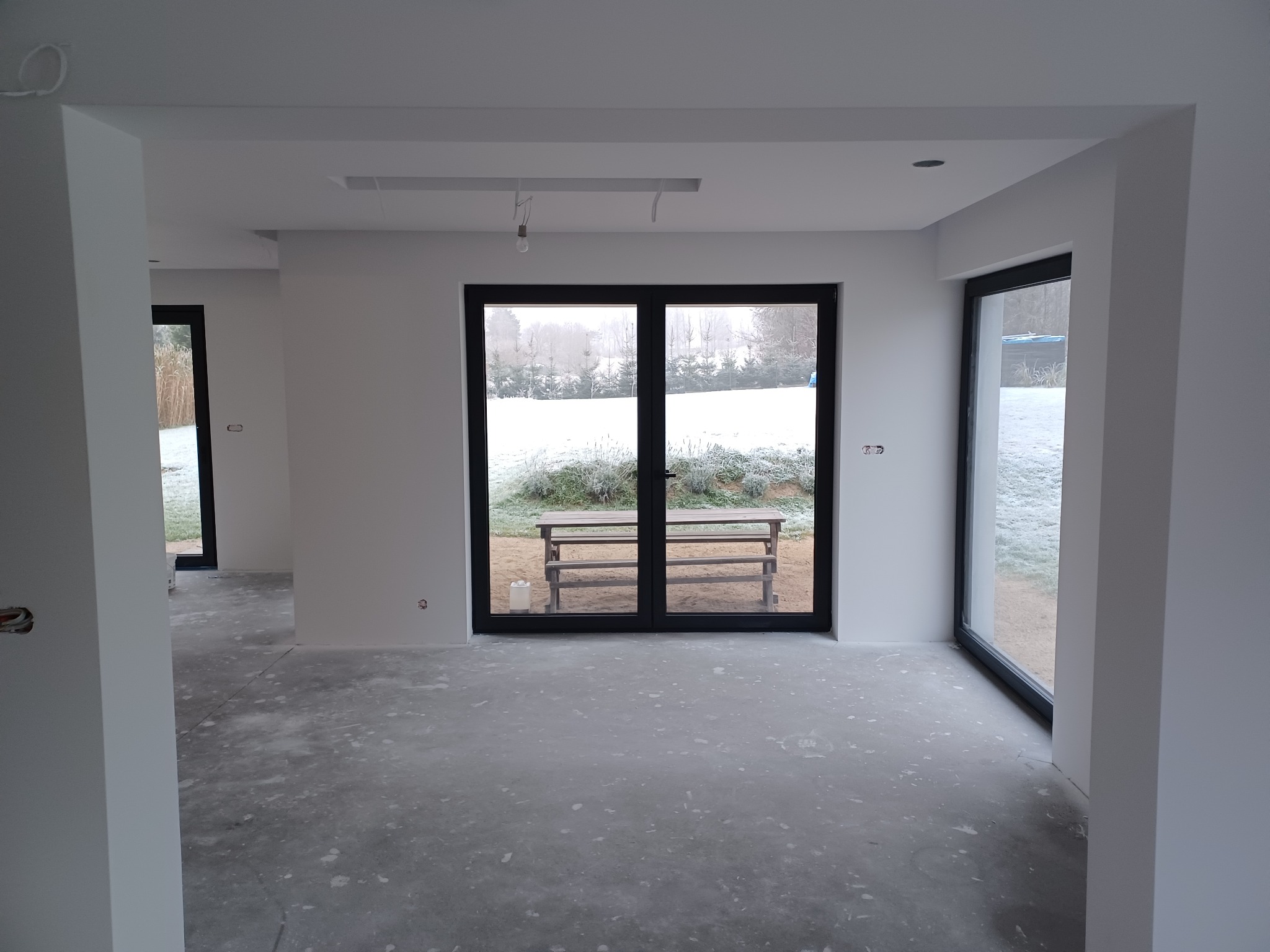Wnętrze w trakcie wykańczania: świeżo pomalowane ściany na biało, surowa podłoga, duże okna z widokiem na zimowy ogród. Widoczne niedokończone elementy sufitu.