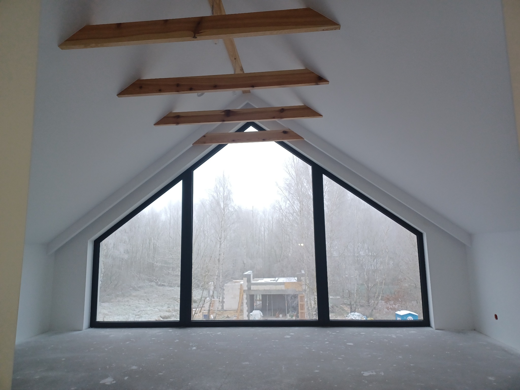 Poddasze z oknem w kształcie trapezu, wykończone gładzią, z widocznymi drewnianymi elementami konstrukcyjnymi pod sufitem. Świeżo wykończone ściany i podłoga.