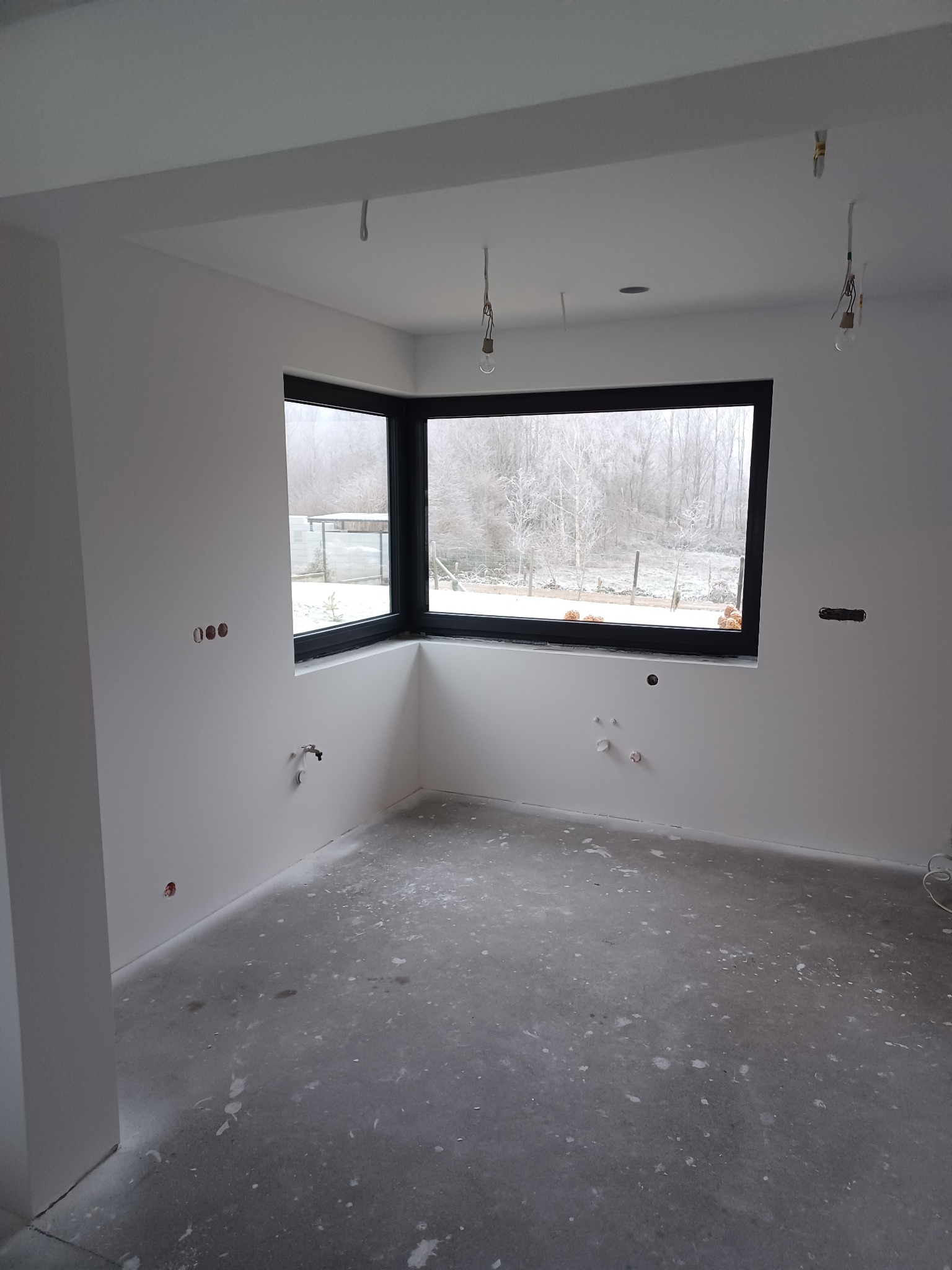 Wnętrze po szpachlowaniu ścian i sufitu, z widocznymi instalacjami elektrycznymi i hydraulicznymi. Duże okno z czarną ramą wychodzi na zimowy krajobraz.