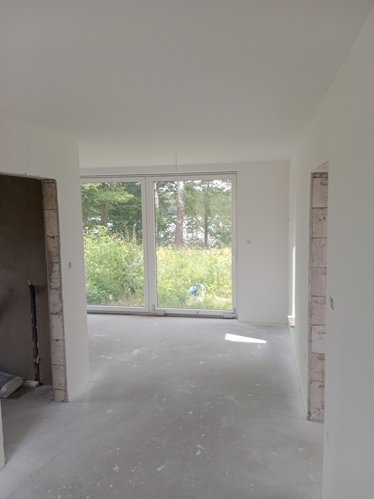 Wnętrze w trakcie prac wykończeniowych: surowe ściany, wylewka podłogowa, widok na zieleń przez okno. Przygotowanie do dalszych etapów remontu.