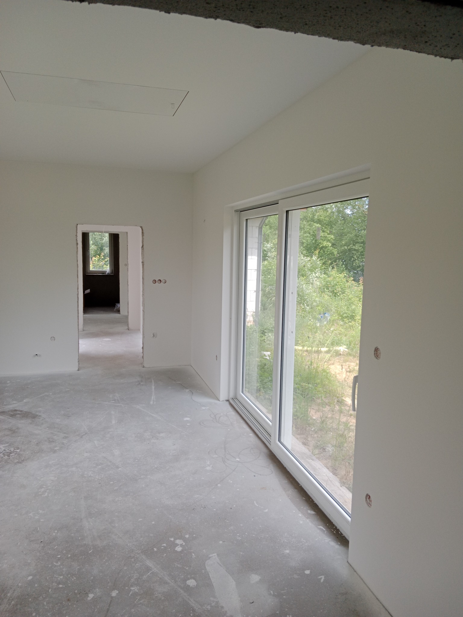 Wnętrze w stanie deweloperskim z gładkimi, białymi ścianami i sufitami. Widoczne okno przesuwne z widokiem na zieleń, surowa podłoga i otwór drzwiowy do kolejnego pomieszczenia.