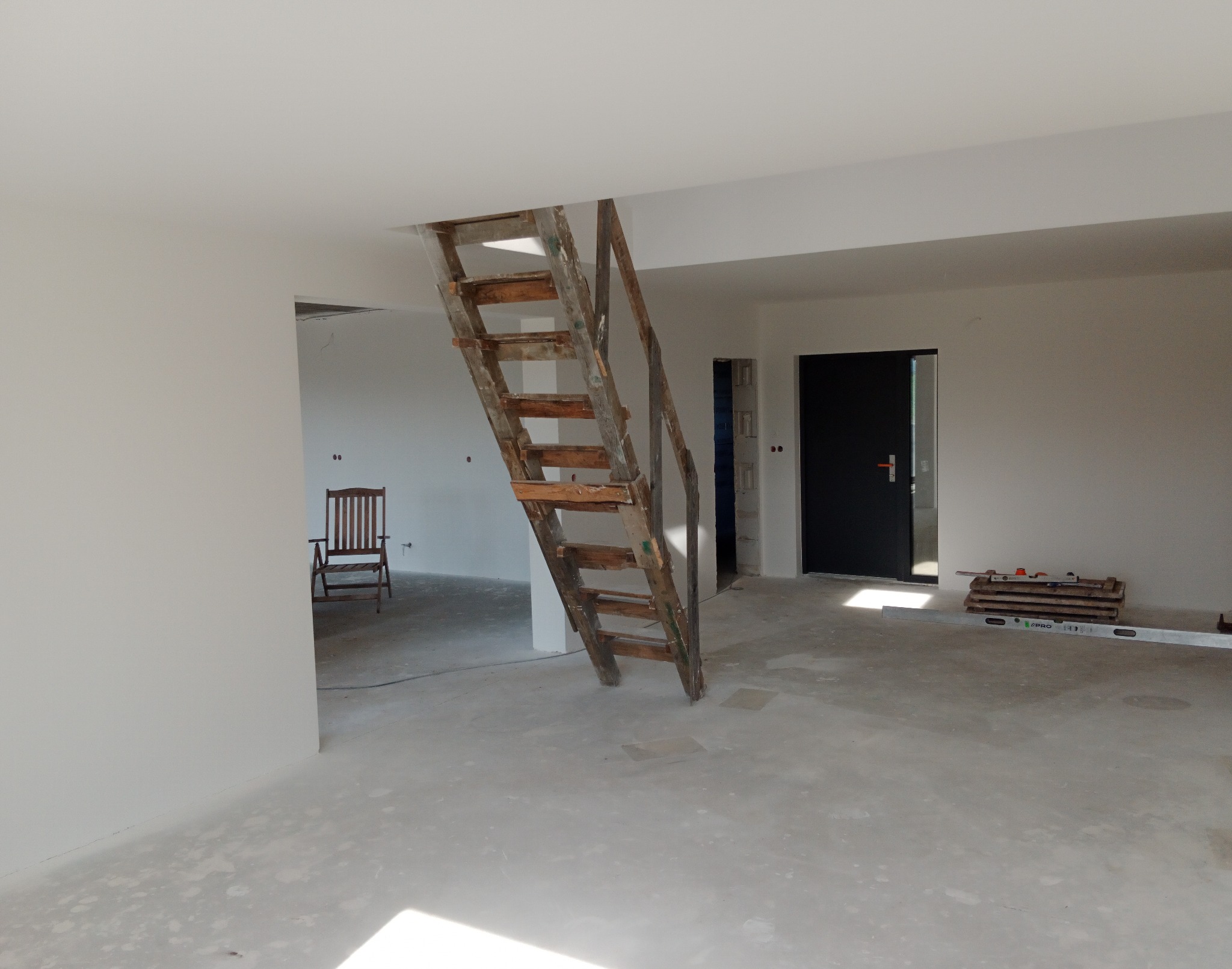 Wnętrze w trakcie remontu: białe ściany, surowy beton na podłodze, drewniana drabina oparta o otwór w stropie, krzesło w tle i narzędzia przy drzwiach.