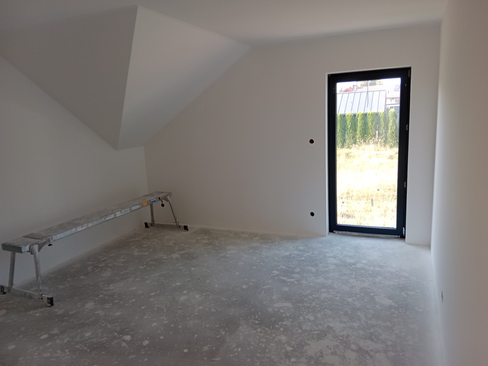 Poddasze po szpachlowaniu i malowaniu na biało, widoczny aluminiowy podest malarski i okno z czarną ramą. Ściany gładkie, przygotowane do dalszych prac wykończeniowych.