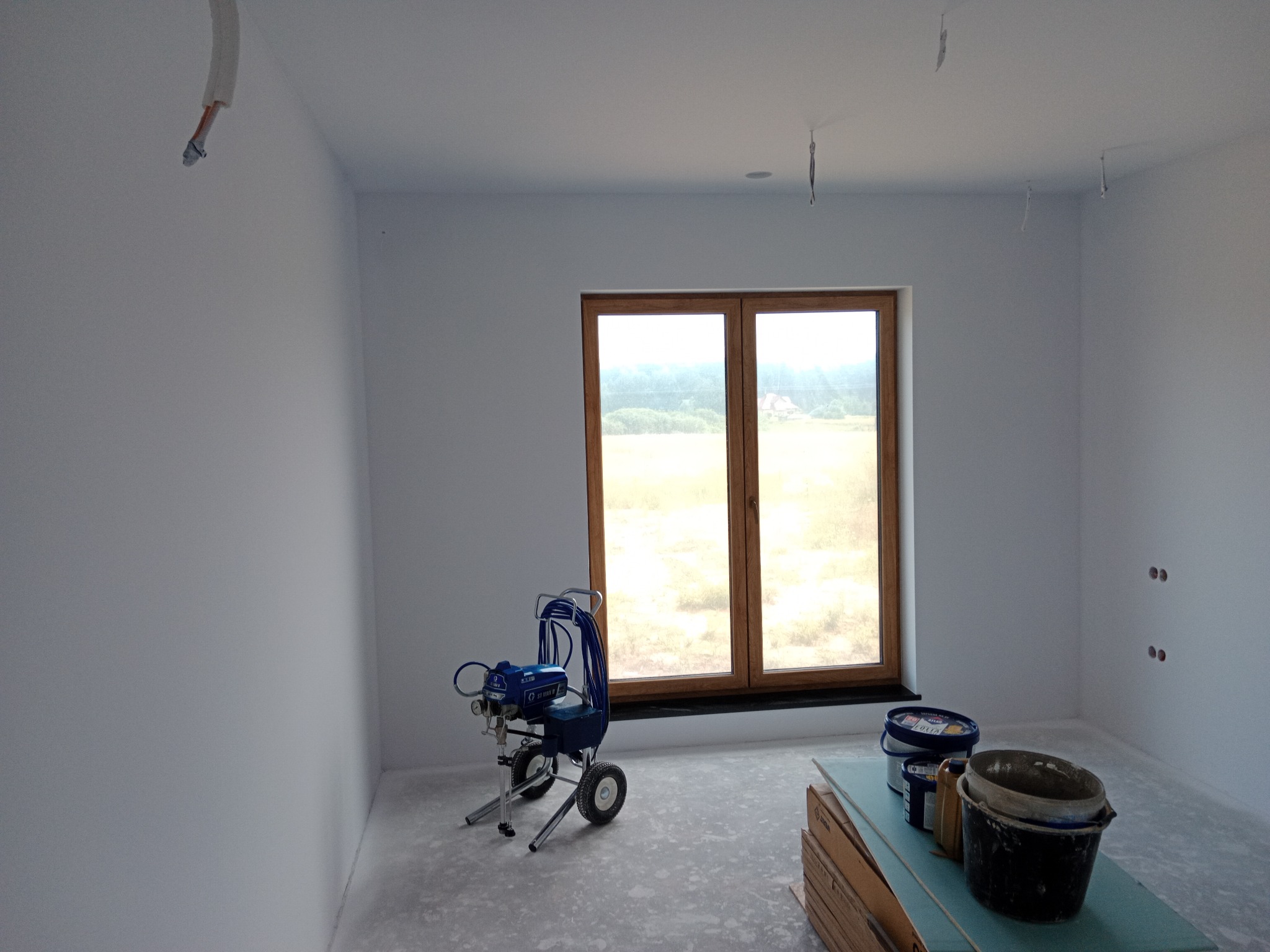Wnętrze po malowaniu natryskowym: białe ściany, okno z drewnianą ramą, agregat malarski i akcesoria malarskie. Widoczne ślady farby na podłodze.