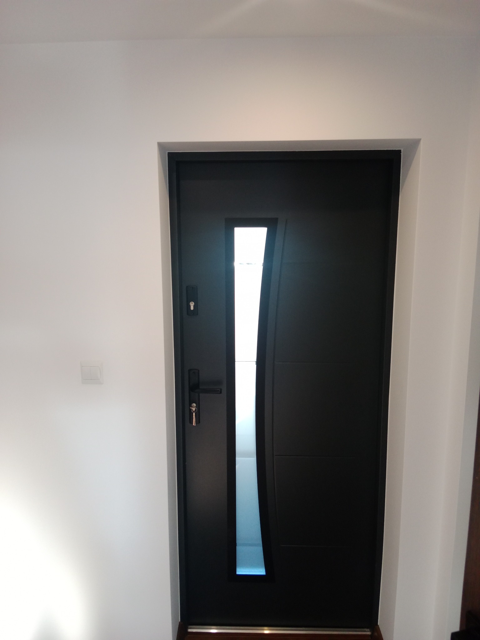 Nowoczesne, czarne drzwi wejściowe z przeszkleniem w kształcie fali, zamontowane w białej ścianie. Widoczny fragment włącznika światła.