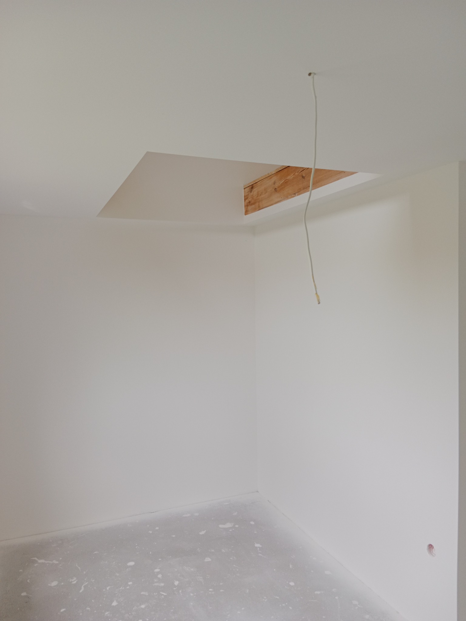 Wnętrze pokoju po remoncie: świeżo pomalowane ściany na biało, widoczny fragment drewnianej konstrukcji w suficie i wystający kabel. Podłoga z drobnymi zabrudzeniami.