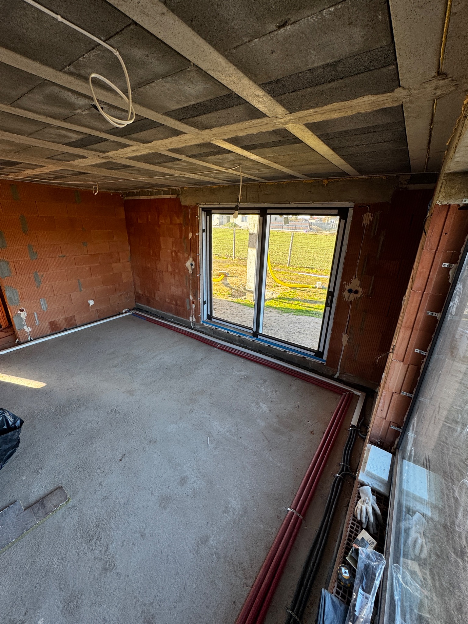 Surowy stan wnętrza budynku w trakcie prac instalacyjnych: widoczne przewody elektryczne pod sufitem, rury przy podłodze, ściany z cegieł dziurawki, okno z widokiem na zewnątrz.