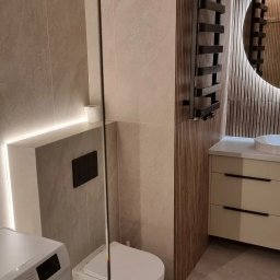 Probud Piotr Rosinski - Nowoczesna łazienka z beżowymi płytkami, podświetleniem LED, wiszącą toaletą i grzejnikiem drabinkowym na tle drewnianej ściany. Pralka widoczna z lewej strony.