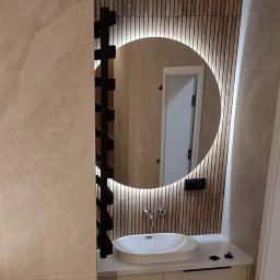 Probud Piotr Rosinski - Nowoczesna łazienka z okrągłym lustrem podświetlanym LED, drewnianymi lamelami na ścianie i minimalistyczną umywalką nablatową. Czarny grzejnik drabinkowy po lewej.