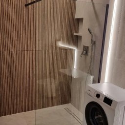 Probud Piotr Rosinski - Nowoczesna łazienka z drewnianą ścianą z lameli, szklaną kabiną prysznicową i subtelnym oświetleniem LED. Uwagę zwraca pralka wkomponowana w aranżację.