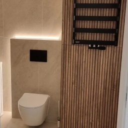Probud Piotr Rosinski - Nowoczesna łazienka z podświetleniem LED, wiszącą toaletą i drewnianą ścianką z grzejnikiem drabinkowym. Minimalistyczny design, beżowe płytki i czarne akcenty.