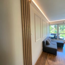 Probud Piotr Rosinski - Nowoczesne wykończenie wnętrza: drewniane lamele na ścianie, listwy dekoracyjne i subtelne oświetlenie LED. Minimalistyczny design z szarą sofą i parkietem.