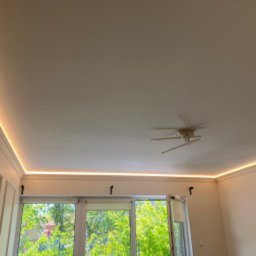 Probud Piotr Rosinski - Jasne wnętrze z oknem i subtelnym oświetleniem LED podsufitowym, podkreślającym architekturę pomieszczenia. Widoczny fragment sufitu z wentylatorem.