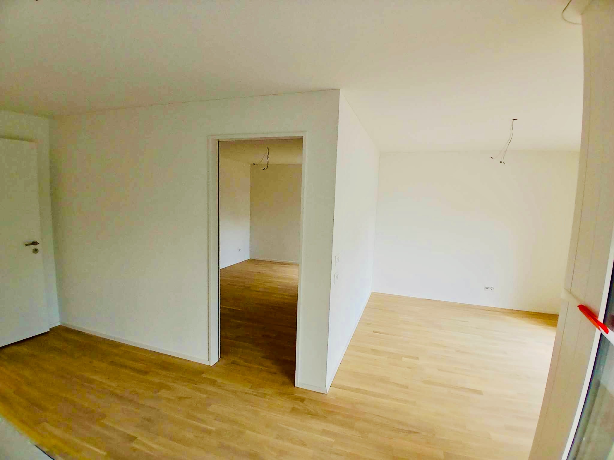 Pusty, świeżo wykończony pokój z jasnymi ścianami i parkietem; widoczne wystające przewody elektryczne w suficie sugerują prace instalacyjne w toku.