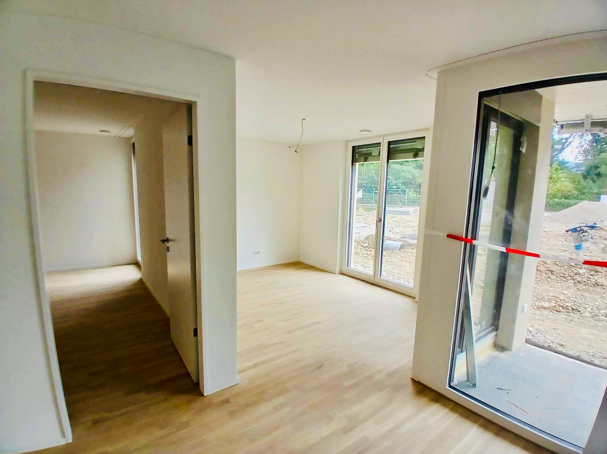 Puste, świeżo wykończone wnętrze z jasnymi ścianami, parkietem i dużymi oknami balkonowymi z widokiem na plac budowy, widoczne kable wystające z sufitu.