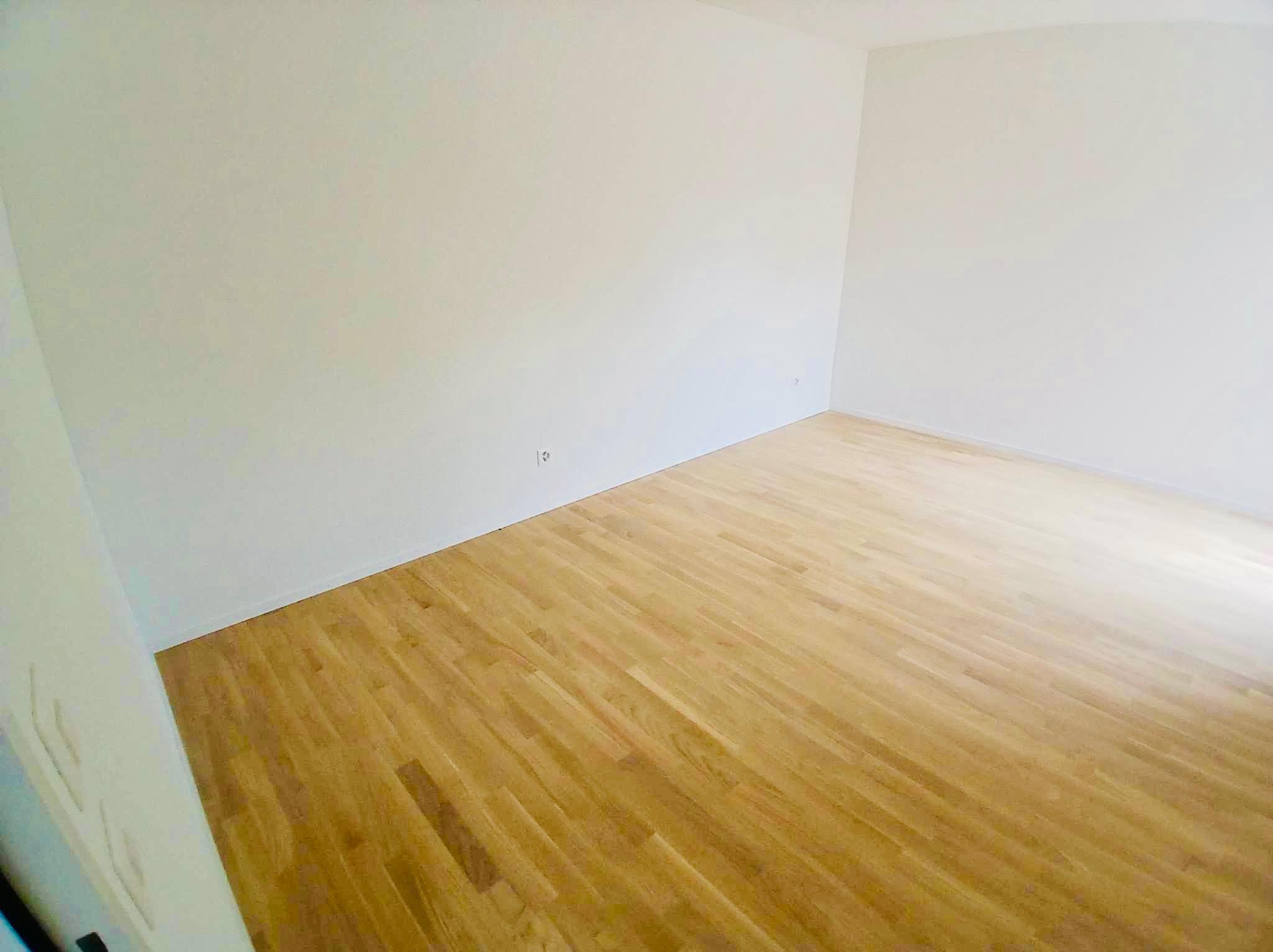 Pusty, świeżo wyremontowany pokój z parkietem w odcieniach brązu i beżowymi ścianami, widoczne dwa gniazdka elektryczne na ścianie.