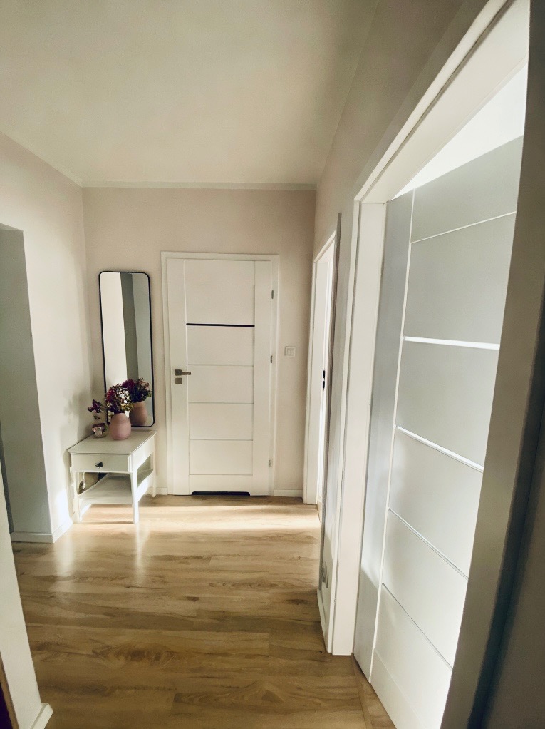 Jasny korytarz z drewnianą podłogą, białymi drzwiami z poziomymi wstawkami, lustrem w czarnej ramie i małym białym stolikiem z kwiatami w wazonie.