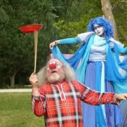 Klaun z czerwonym nosem w kraciastej koszuli balansuje talerzem na patyku, obok kobieta w niebieskim kostiumie i peruce na tle zieleni.