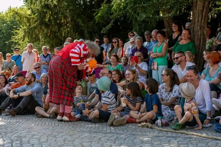 Klaun w czerwono-białym stroju i kraciastej spódnicy rozmawia z dziećmi siedzącymi na ziemi, w tle tłum widzów pod drzewami.