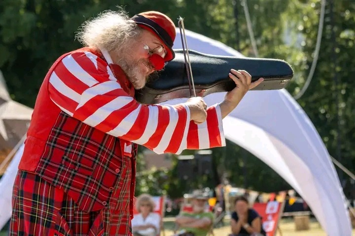 Klaun w czerwono-białym stroju i czerwonym nosie, trzymający futerał na skrzypce udając grę, na tle białego namiotu i publiczności.