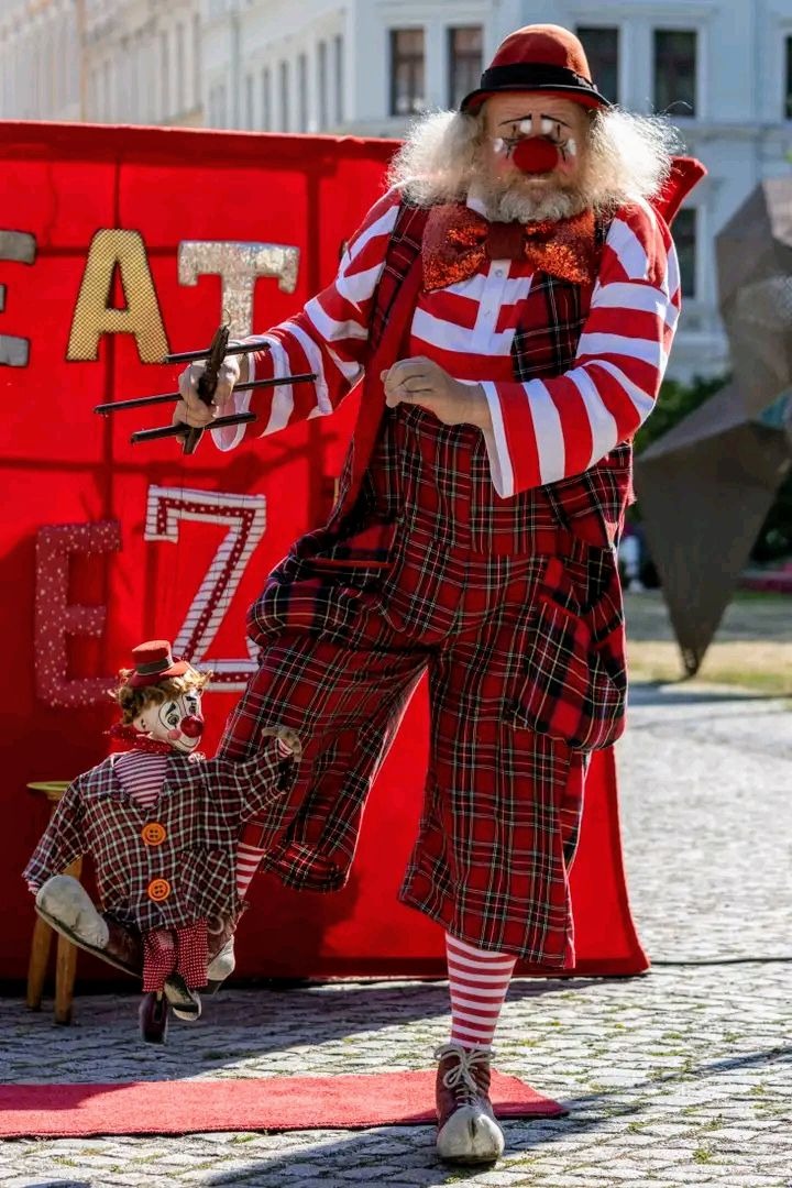 Klaun w czerwono-białym stroju w kratę, z lalką klauna, prezentuje sztuczkę z czarnymi prętami na tle czerwonego baneru.