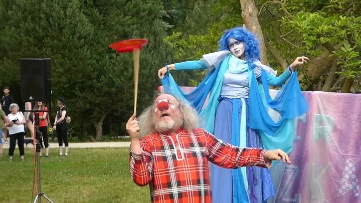 Klaun z czerwonym nosem w kraciastej koszuli balansuje talerzem na patyku, obok kobieta w niebieskim kostiumie i peruce na tle zieleni.