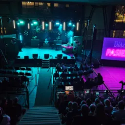 Widok z góry na scenę podczas wydarzenia 'Disco Pasieka', z publicznością siedzącą na trybunach, oświetlenie sceniczne w odcieniach turkusu i różu, oraz balon z logo wydarzenia po lewej stronie.