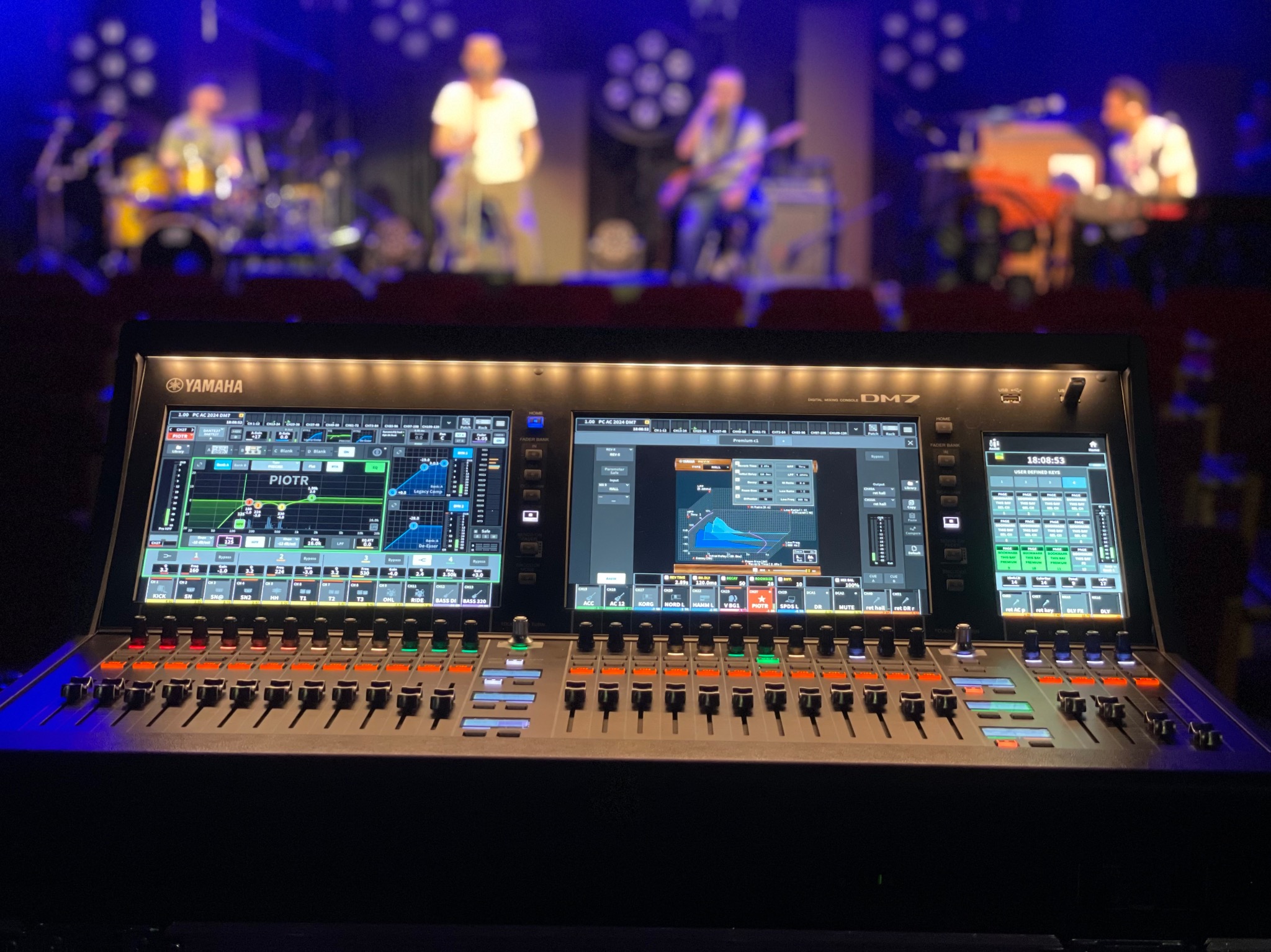 Profesjonalny mikser Yamaha DM7 z podświetlanymi suwakami i ekranami dotykowymi, ustawiony przed sceną z zespołem muzycznym w tle, oświetlonym reflektorami.