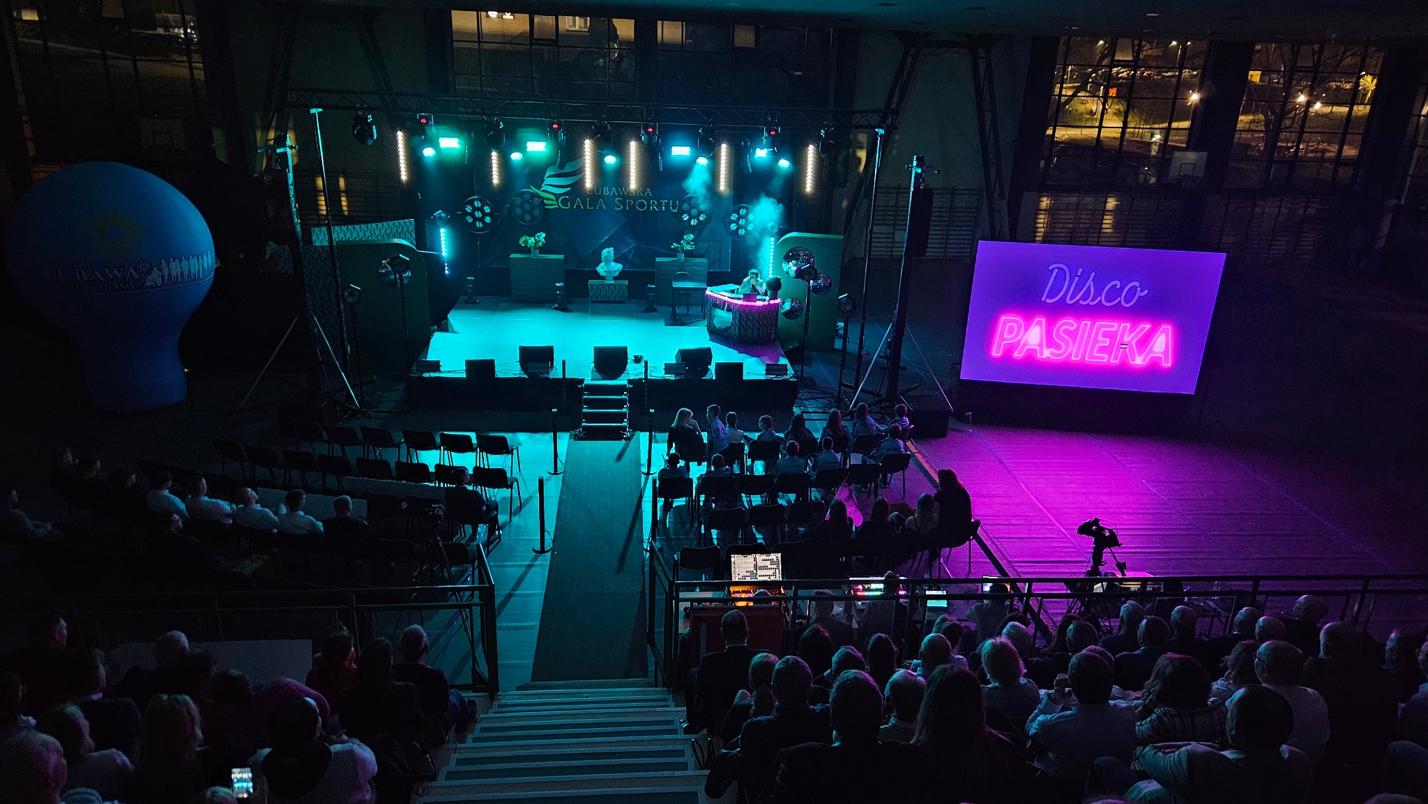 Widok z góry na scenę podczas wydarzenia 'Disco Pasieka', z publicznością siedzącą na trybunach, oświetlenie sceniczne w odcieniach turkusu i różu, oraz balon z logo wydarzenia po lewej stronie.