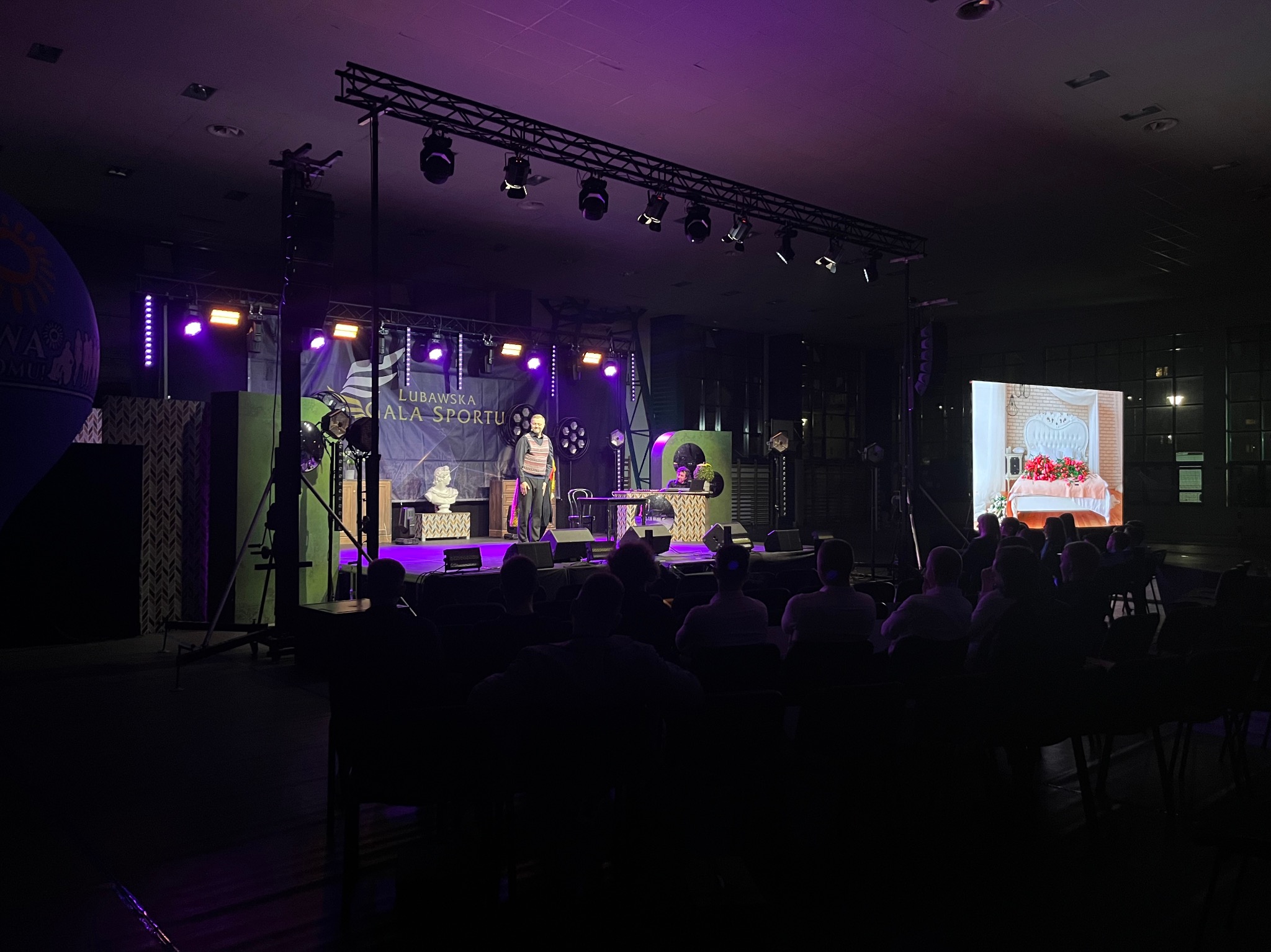 Scena oświetlona fioletowymi światłami podczas wydarzenia w Gdyni, widoczny prelegent na tle dekoracji i publiczność na widowni.