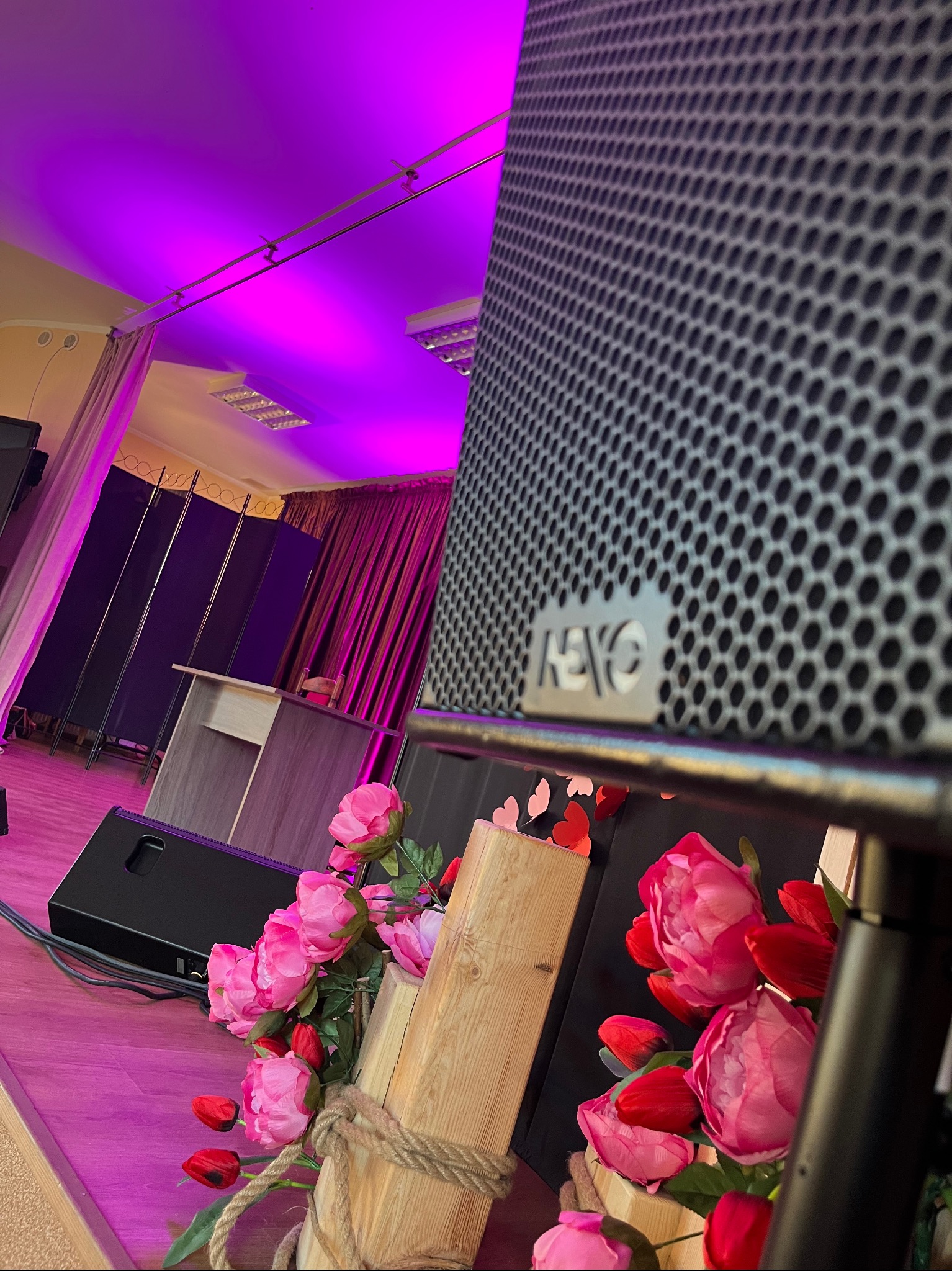 Duży głośnik marki RCF z metalową siatką na tle sceny oświetlonej fioletowym światłem, z dekoracją z różowych i czerwonych kwiatów w drewnianej oprawie ze sznurem.