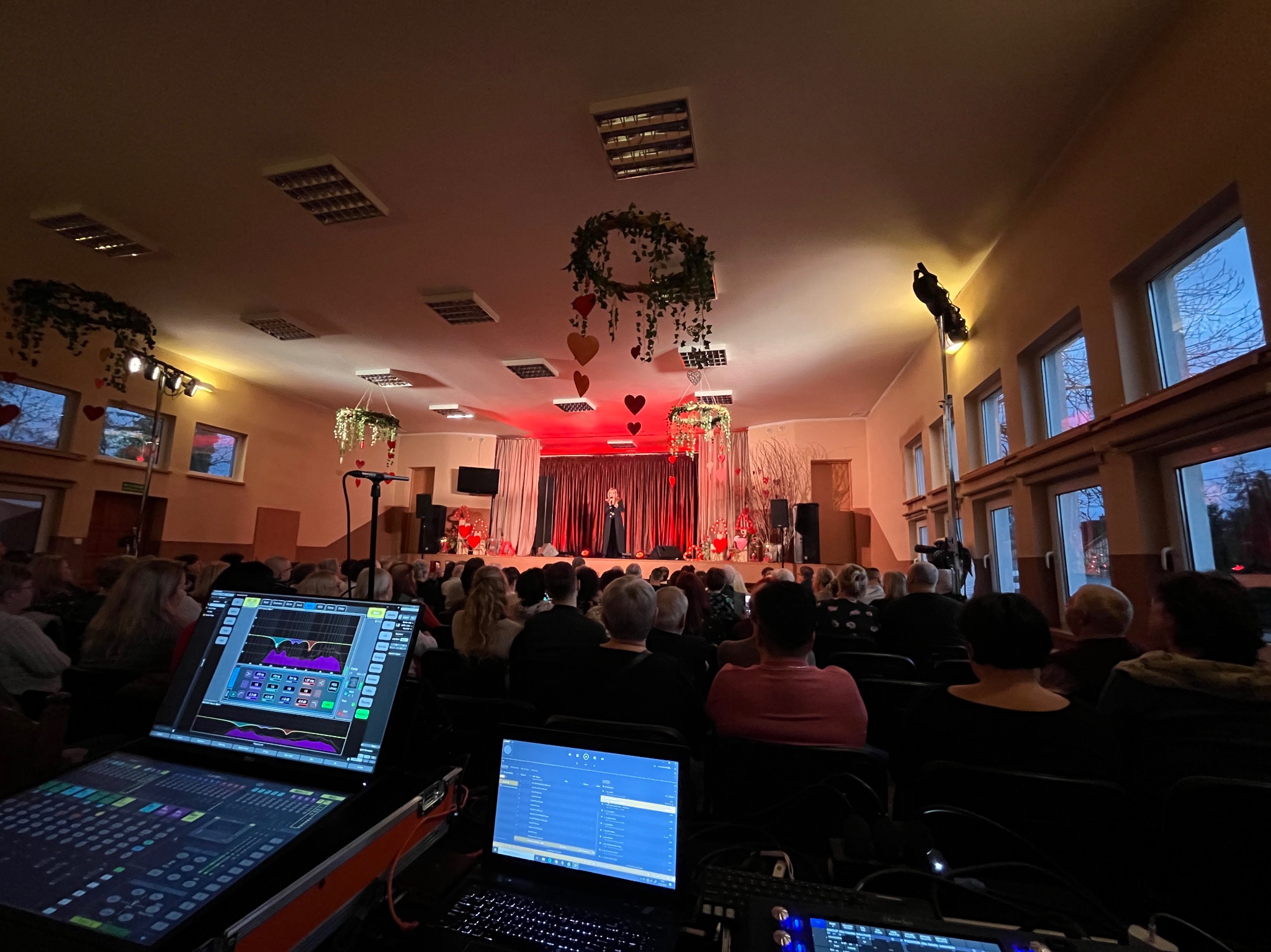 Widok zza konsolety dźwiękowej na scenę oświetloną czerwonym światłem, gdzie występuje osoba przed publicznością, dekoracje z sercami, sala z oknami w tle.