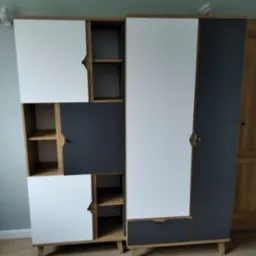 Dwa zestawy nowoczesnych mebli modułowych w kolorach bieli i grafitu, z drewnianymi elementami i nóżkami, ustawione na podłodze imitującej drewno, na tle zielonej ściany.