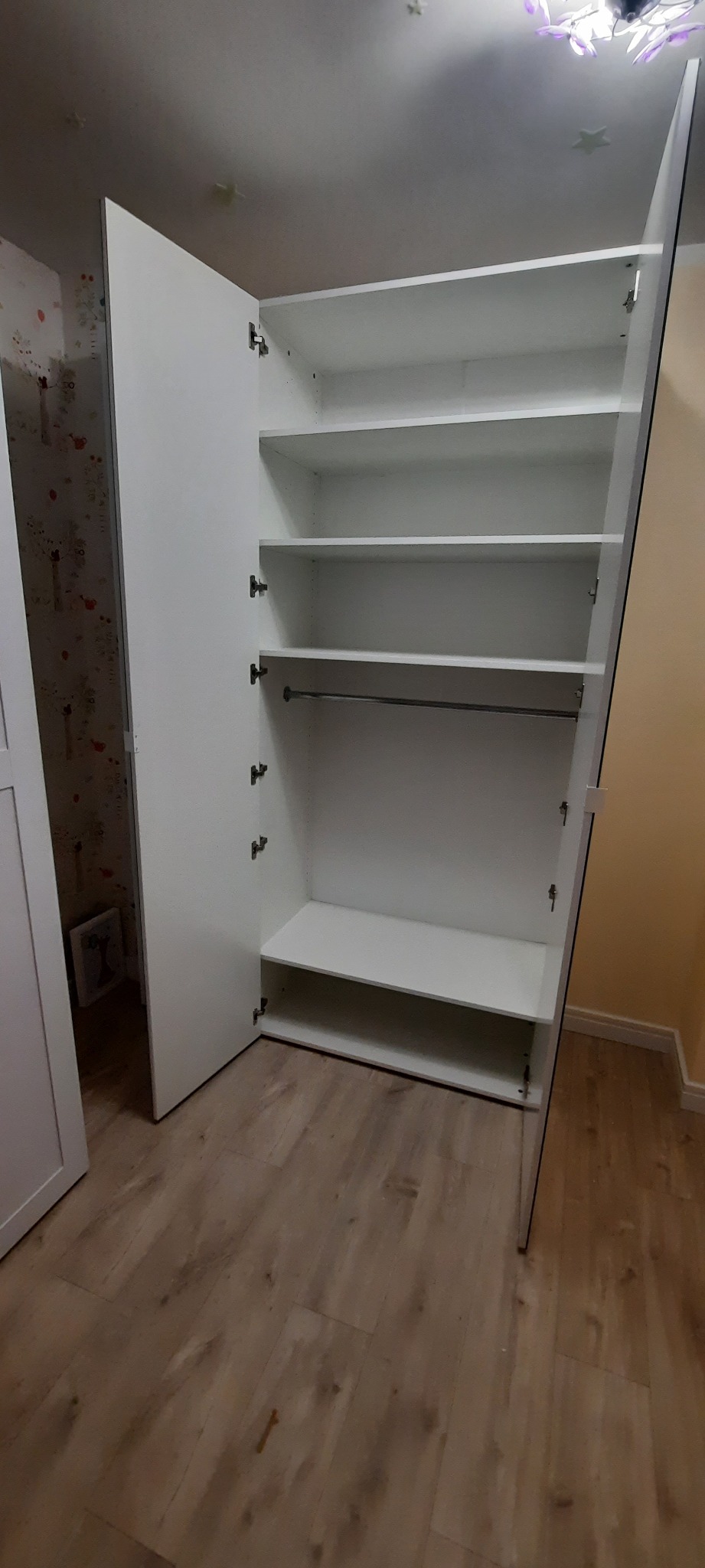 Biała, otwarta szafa wnękowa z półkami i drążkiem na ubrania, zamontowana w mieszkaniu w Słupsku. Widoczne drewniane panele podłogowe.