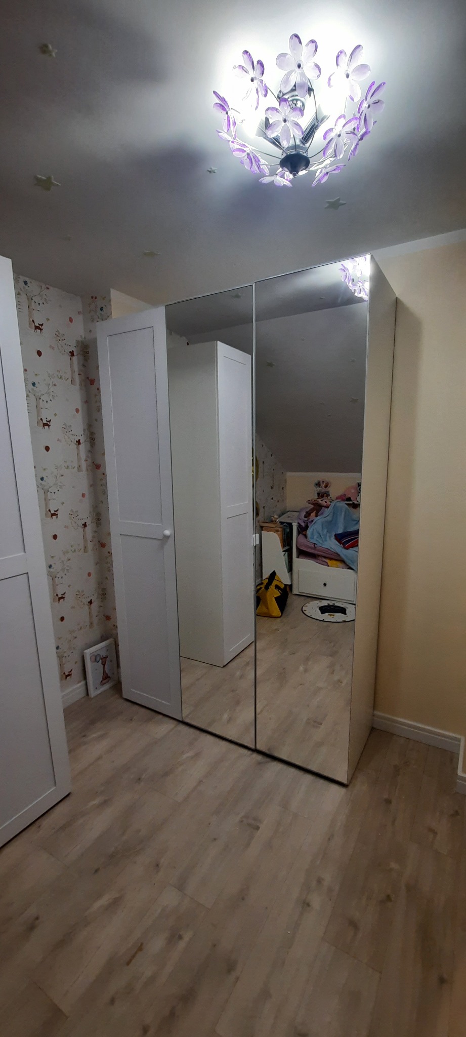Trzydrzwiowa szafa z lustrzanymi frontami w pokoju dziecięcym, widoczna na tle ściany z tapetą w zwierzątka i lampy w kształcie kwiatów na suficie.