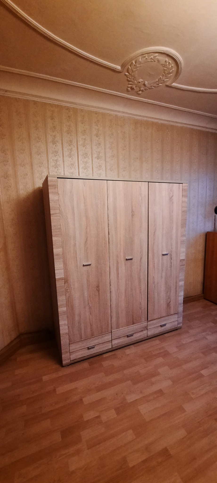 Trzydrzwiowa szafa z drewnopodobnym fornirem, z trzema szufladami na dole, na tle tapety w pionowe pasy i drewnianej podłogi.