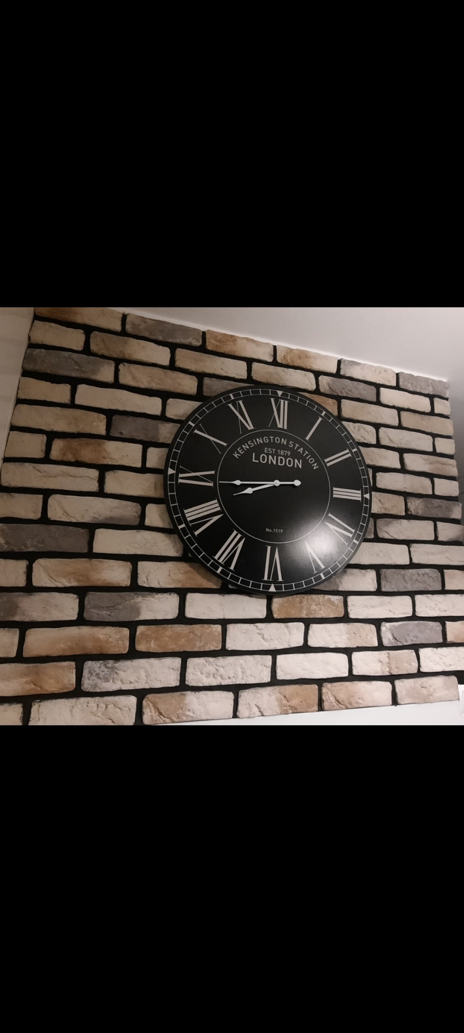 Ściana wyłożona dekoracyjną cegłą w odcieniach beżu i szarości z dużym, okrągłym zegarem w stylu retro wskazującym godzinę.