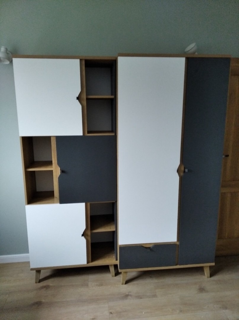 Dwa zestawy nowoczesnych mebli modułowych w kolorach bieli i grafitu, z drewnianymi elementami i nóżkami, ustawione na podłodze imitującej drewno, na tle zielonej ściany.