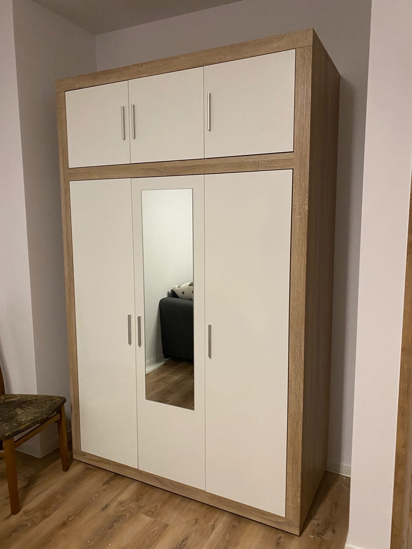 Szafa trzydrzwiowa z lustrem pośrodku, w odcieniach bieli i drewna, z dodatkową nadstawką, stojąca na panelach podłogowych, odbicie sofy w lustrze.