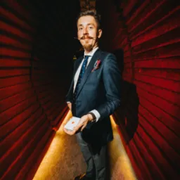 Mężczyzna w garniturze z wąsami trzymający talię kart na tle czerwonych draperii i oświetlonej podłogi.