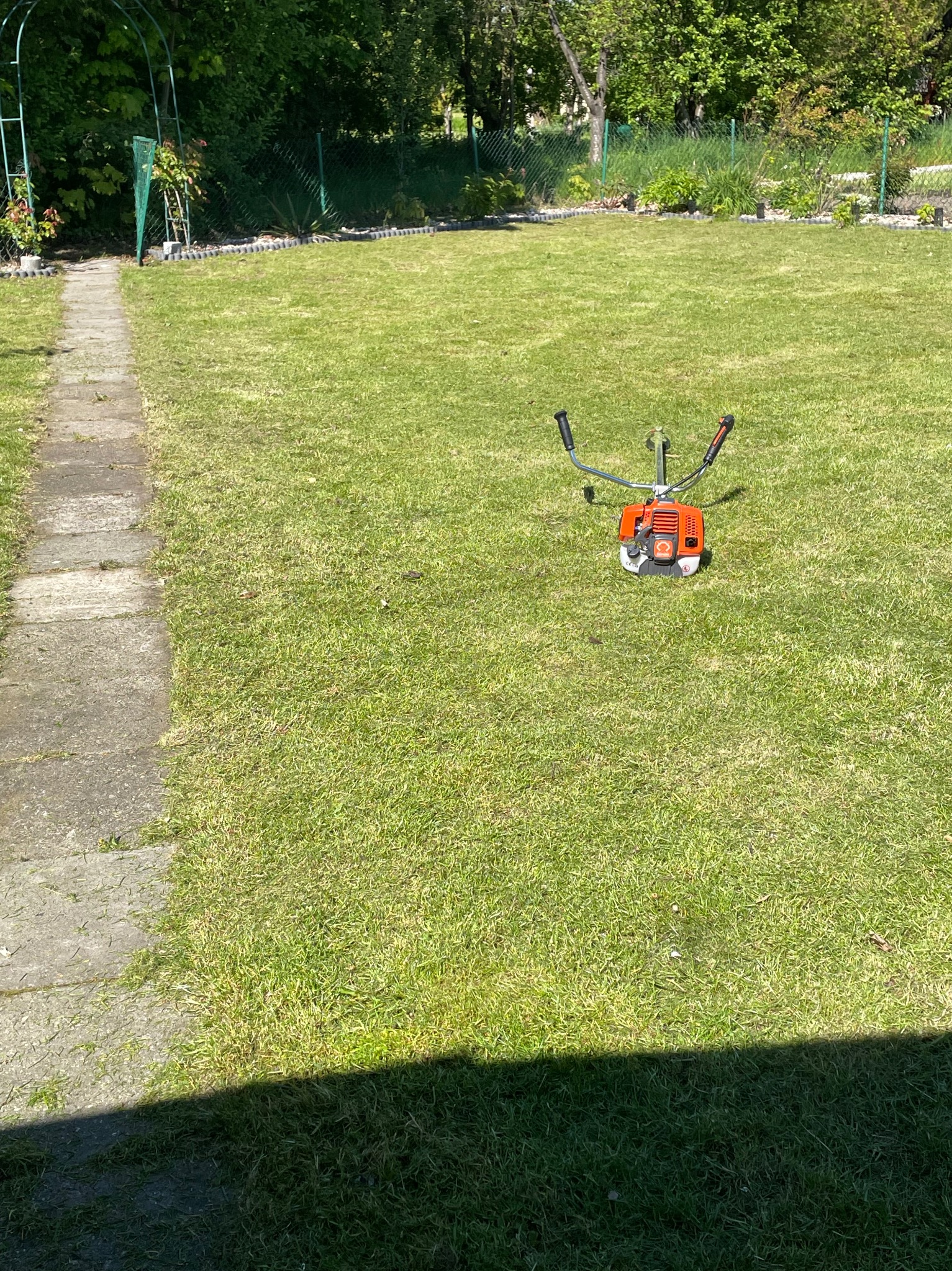 Pomarańczowa kosiarka spalinowa leży na trawniku w słoneczny dzień, widoczny fragment kamiennej ścieżki i zielona pergola w tle.