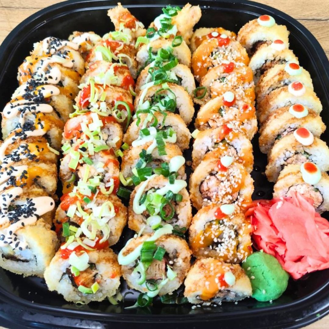 Czarny talerz z różnorodnym zestawem sushi: rolki z tempurą, posypane sezamem, polane sosem teriyaki, udekorowane szczypiorkiem i imbirem.