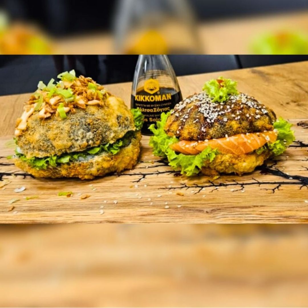 Dwa nietypowe burgery z ryby i ryżu w tempurze, podane na drewnianej desce z butelką sosu sojowego Kikkoman w tle.